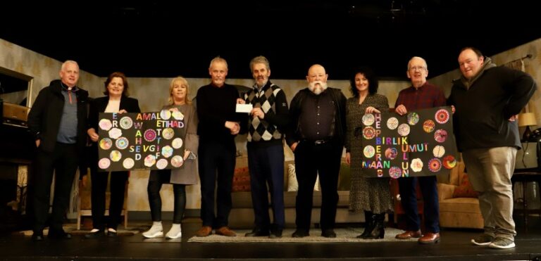 A man presents a cheque to a Trocaire representative, along with 7 people holding artwork made by childeren