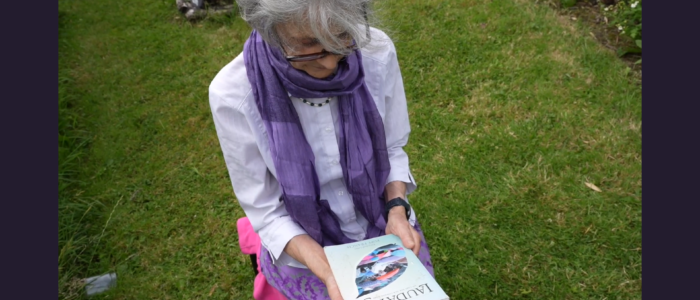 Screenshot 2025-09-01 134506 Picture of a woman in a purple scarf and a white shirt kneeling down in a garden with a book.